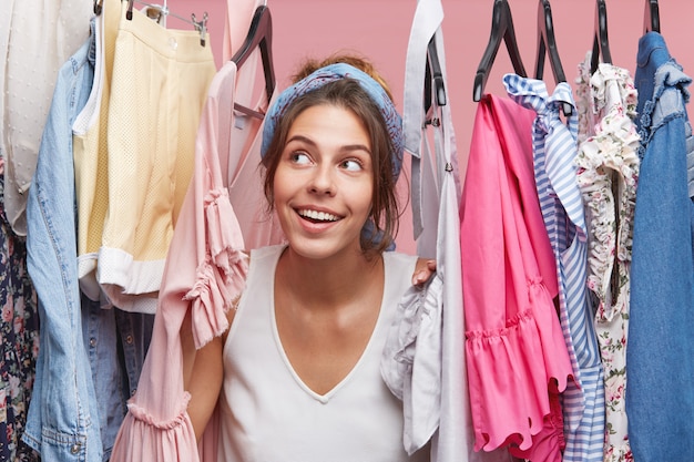 Nette Frau mit verträumtem Ausdruck, der durch Kleiderbügel mit Kleidern schaut und von neuem modischem Kleid oder Bluse träumt. Entzückende Frau, die davon träumt, am Wochenende mit Freunden einkaufen zu gehen