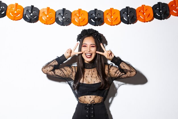 Nette glückliche asiatische Frau, die Halloween-Partei genießt, Friedensgeste zeigt und lächelt, Hexenkostüm tragend, gegen Kürbisfahnendekoration stehend.