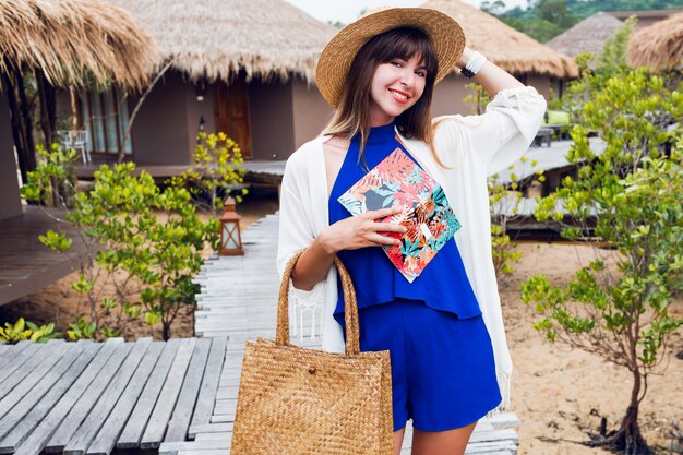 Nette glückliche reisende Frau mit dem lächelnden und schauenden Notizbuch. Blauer Overall, Strohhut und Tasche, Sonnenbrille. Brünettes Mädchen, das in ihrer erstaunlichen Luxusvilla aufwirft.