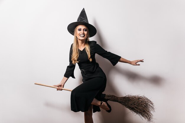 Nette Hexe im langen Kleid sitzt auf Besen mit Lächeln. Sorglose blonde Dame im Karnevalskostüm, die Halloween genießt.
