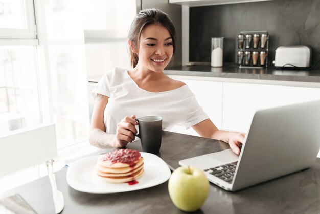 Nette junge Frau, die an der Küche unter Verwendung des Laptops sitzt