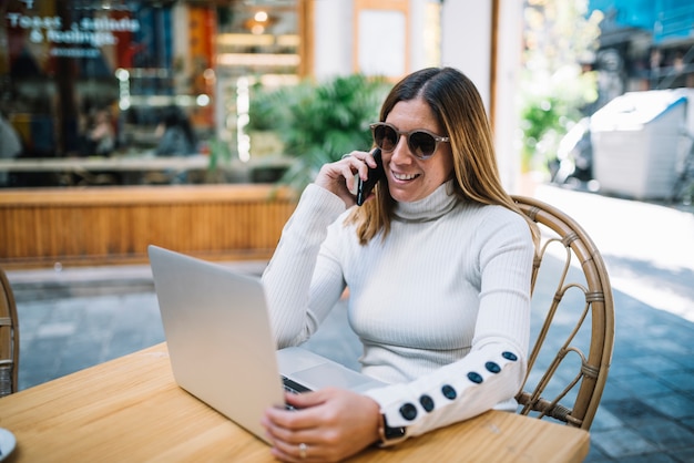 Nette junge Frau, die bei Tisch Laptop und Smartphone im Straßencafé verwendet