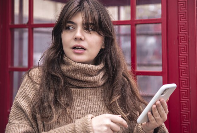 Nette junge Frau mit einem Smartphone auf dem Hintergrund einer roten Telefonzelle.