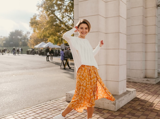 Nette junge glückliche lächelnde Frau in gelbem bedrucktem Kleid und gestricktem weißem Pullover an einem sonnigen Herbsttag