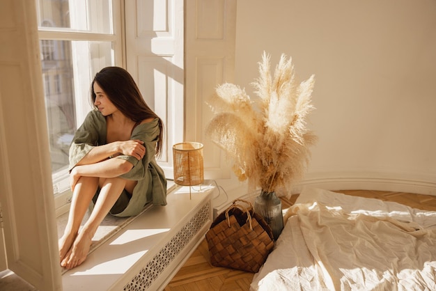 Nette junge kaukasische Frau blickt aus dem Fenster, während sie auf der Fensterbank sitzt und das schöne Wetter genießt. Brunette trägt lässige Sommerkleidung zu Hause Stilvolles Urlaubskonzept