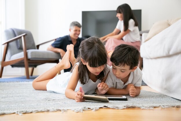 Nette kleine Kinder, die auf Boden im Wohnzimmer liegen und digitale Geräte mit Lern-Apps verwenden, während Eltern lachen