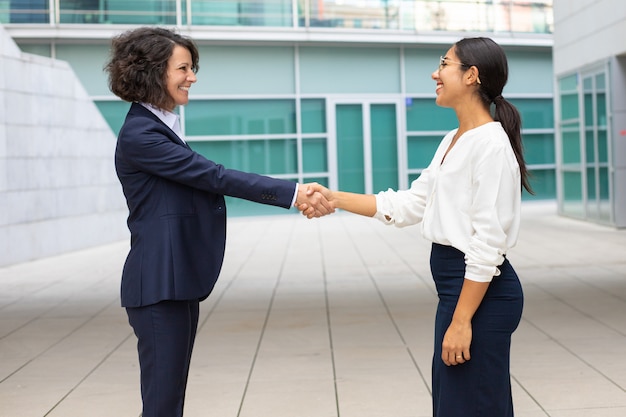 Nette Kollegen, die Hände nahe Bürogebäude rütteln. Junge Frauen, welche das Gesellschaftsanzugtreffen im Freien tragen. Business-Handshake-Konzept