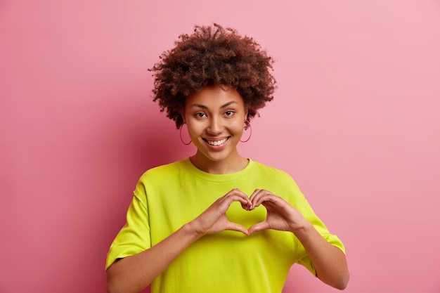 Nette lächelnde afroamerikanische Frau macht ich liebe dich Geste gesteht in Liebe drückt Sympathie aus zeigt Herzzeichen gekleidet in Freizeitkleidung isoliert über rosa Wand