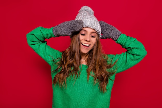 Nette reizende glückliche Frau mit gekleidetem Wintermütze des langen Haares und grünem Pullover, der über rotem Hintergrund mit geschlossenen Augen, glücklichem Lächeln und ruhigen Gefühlen steht