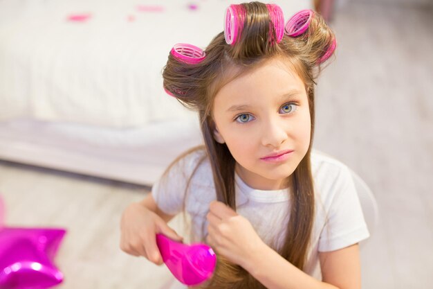 Nette Vorschülerin, die sich um ihr Haar kümmert. Verträumtes kleines Mädchen mit weißem T-Shirt, das ihr Haar mit rosa Haarbürste bürstet und nach oben schaut.