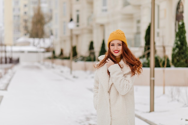 Nette weiße Frau, die im Wintertag aufwirft. Außenfoto der zufriedenen Ingwerdame im langen Mantel.