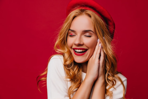 Nette weiße Frau in der französischen Baskenmütze, die Inspiration ausdrückt. Debonair blondes Mädchen lacht mit geschlossenen Augen auf roter Wand.