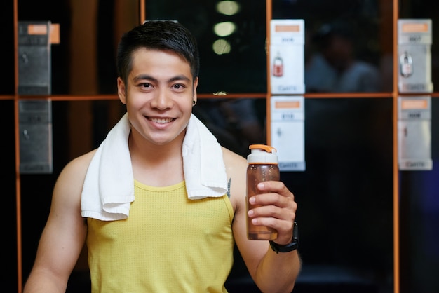 Netter asiatischer Mann, der im Umkleideraum in der Turnhalle mit Sportflasche aufwirft