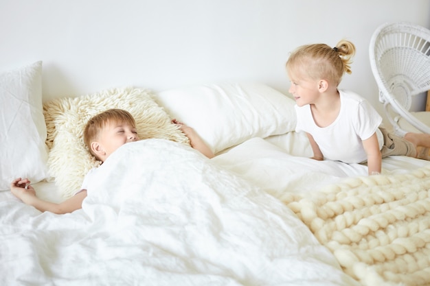 Netter blonder kleiner Junge, der Pyjamas trägt, der auf weißem großem Bett sitzt und seinen älteren Bruder weckt, der neben ihm schläft und Guten Morgen sagt. Zwei Brüder spielen zusammen im Schlafzimmer und haben Spaß