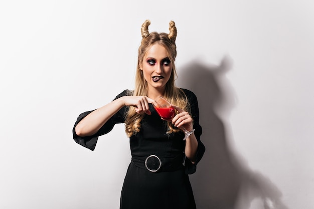 Netter blonder Vampir im Kleid, das auf weißer Wand steht. Hübsches schlankes Mädchen, das Blut in Halloween trinkt.