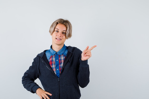 Netter jugendlicher Junge, der auf die obere rechte Ecke im Hemd, im Hoodie zeigt und jovial schaut, Vorderansicht.