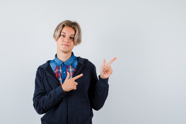 Netter jugendlicher Junge, der auf die obere rechte Ecke im Hemd, im Hoodie zeigt und positiv schaut. Vorderansicht.