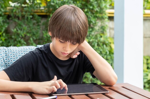 Netter Junge, der Tablette benutzt, während er zu Hause Hausaufgaben im Garten macht