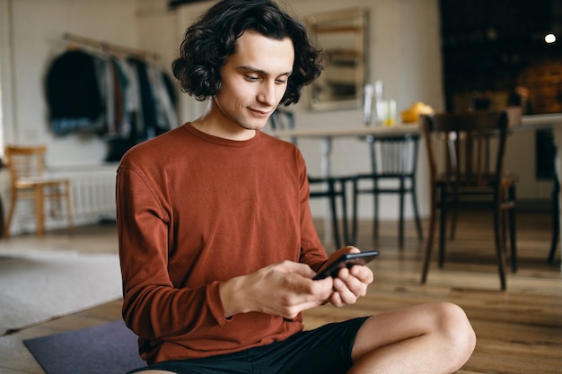 Netter junger männlicher Student mit gewelltem Haar sitzt auf dem Boden zu Hause mit Handy, das Textnachricht an Freunde über Gruppenchat auf Messenger tippt, durch Meme schaut, sein soziales Netzwerkkonto durchsucht