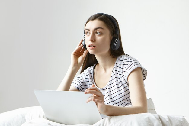 Netter junger weiblicher Übersetzer, der entfernt von zu Hause aus arbeitet und auf einem weißen Bett mit einem tragbaren Computer sitzt