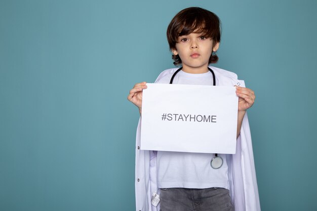 Netter kleiner entzückender Junge im weißen medizinischen Anzug und in den grauen Jeans mit Hashtag des zu Hause bleiben gegen Coronavirus auf blauem Schreibtisch