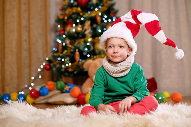 Netter kleiner Junge in Santa's Hut mit Weihnachtsbaum im Hintergrund.