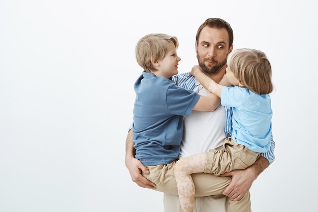 Netter lustiger kaukasischer Vater, der zwei junge Söhne in den Armen hält, jüngeren Jungen anstarrt und Gesichter macht, positiv und glücklich ist, während er großartige Kinder hat