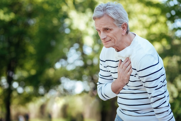 Netter ruhiger älterer mann, der steht und unter schmerzen in seiner brust leidet, während er durch den park geht