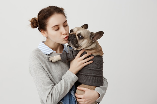 Netter schuss des erwachsenen mädchens, das niedlichen kleinen hund küsst, während es ihn mit zärtlichkeit hält. porträt des hundes und seiner besitzerin, die zeit miteinander kuscheln und freunde sind. anzeige der zuneigung