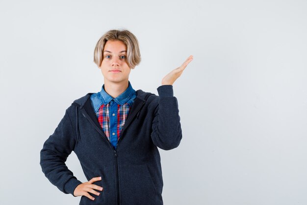 Netter Teenager, der die Palme in Hemd, Hoodie hebt und selbstbewusst aussieht. Vorderansicht.