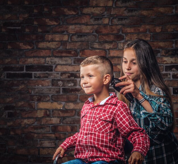 Netter Vorschuljunge, der Haarschnitt bekommt. Die ältere Schwester schneidet ihren kleinen Bruder mit einem Trimmer gegen eine Ziegelwand.