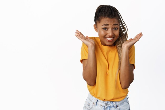 Nettes afrikanisch-amerikanisches Mädchen, das albern zuckt und lächelt, oops, schuldiger Blick, der in gelbem T-Shirt auf weißem Hintergrund steht