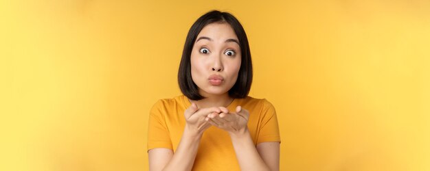Nettes asiatisches Mädchen, das einen Luftkuss sendet, der mwah mit gespitzten Lippen bläst, die über gelbem Hintergrund stehen