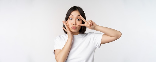 Nettes asiatisches Mädchen, das mit kawaii Vsign-Friedensgeste nahe dem Gesicht aufwirft, das im T-Shirt über weißem Hintergrund steht