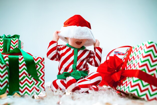 Nettes Baby 1 Jahr alt, das Weihnachtsmütze trägt, der über Weihnachtsdekorationen mit Geschenken aufwirft. Mit Weihnachtskugel auf dem Boden sitzen. Ferienzeit.
