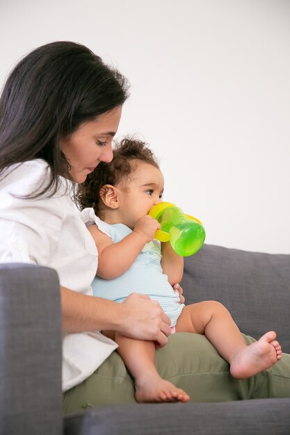 Nettes Baby, das auf Mutterschoß sitzt und Wasser von Flasche trinkt. Vertikaler Schuss. Elternschafts- und Kindheitskonzept