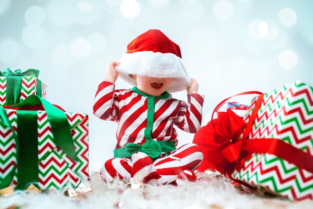 Nettes Baby, das Weihnachtsmütze trägt, die über Weihnachtsdekorationen mit Geschenken aufwirft. Mit Weihnachtskugel auf dem Boden sitzen. Ferienzeit.