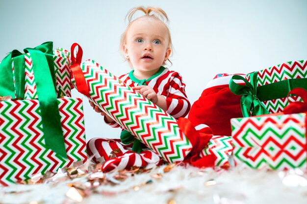 Nettes Baby nahe der Weihnachtsmannmütze, die über Weihnachtshintergrund mit Dekoration aufwirft. Mit Weihnachtskugel auf dem Boden sitzen. Ferienzeit.