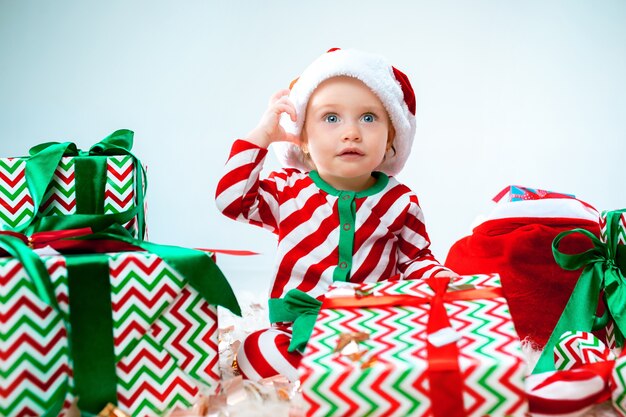 Nettes Baby nahe der Weihnachtsmannmütze, die über Weihnachtshintergrund mit Dekoration aufwirft. Mit Weihnachtskugel auf dem Boden sitzen.