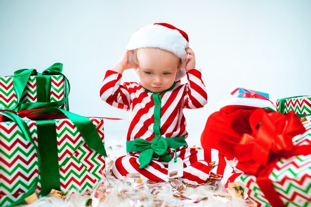 Nettes Baby nahe der Weihnachtsmannmütze, die über Weihnachtshintergrund mit Dekoration aufwirft. Mit Weihnachtskugel auf dem Boden sitzen.