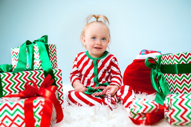 Nettes Baby nahe der Weihnachtsmannmütze, die über Weihnachtshintergrund mit Dekoration aufwirft. Mit Weihnachtskugel auf dem Boden sitzen.