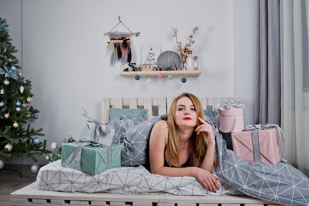 Nettes blondes Mädchen auf schwarzem BH, das auf dem Bett mit Geschenkboxen gegen Neujahrsbaum im Studio liegt Frohe Feiertage