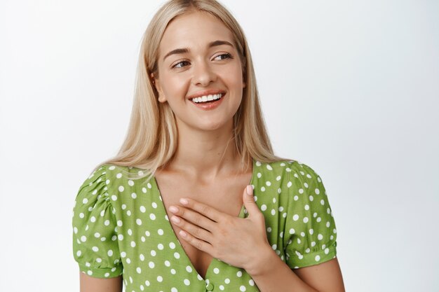 Nettes blondes Mädchen im Kleid, das Hand auf der Brust hält, lächelt und auf Weiß beiseite schaut. Platz kopieren