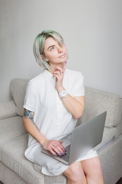 Nettes blondes Mädchen mit kurzer Frisur sitzt auf Sofa im Studio. Sie hat einen Laptop auf den Knien, ein weißes Kleid. Sie träumt.