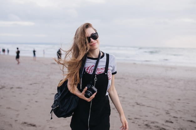 Nettes braunhaariges mädchen mit kamera, die auf natur in bewölktem tag aufwirft. außenaufnahme der gutaussehenden dame mit dem schwarzen rucksack, der um strand geht.