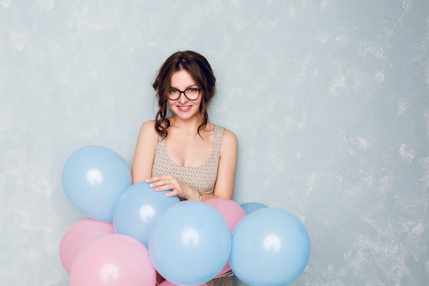 Nettes brünettes Mädchen, das in einem Studio steht, breit lächelt und blaue und rosa Luftballons hält.