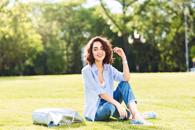 Nettes brünettes Mädchen mit kurzen Haaren, die auf Gras im Park aufwerfen. Sie trägt weißes T-Shirt, Hemd und Jeans, Schuhe. Sie sieht im Sonnenlicht glücklich aus.