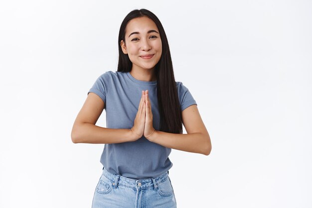 Nettes dummes asiatisches brünettes Mädchen mit zärtlichem Ausdruck, lächelnd und freundlich anstarrender Kamera, hält die Hände im Gebet und bittet um Hilfe, bittet um Hilfe oder fleht etwas an, um Ihnen zu danken, dass Sie dankbar sind