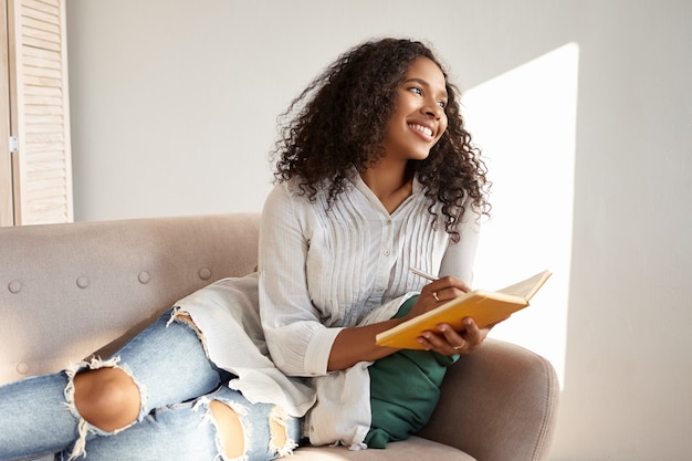 Nettes entzückendes afroamerikanisches Studentenmädchen mit schwarzem voluminösem Haar, das Freizeit nach dem College genießt, auf Couch in stilvollen zerrissenen Jeans und Bluse liegend, Gedanken und Ideen in ihrem Tagebuch teilend