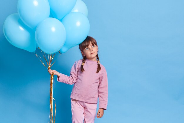 Nettes europäisches weibliches Kind, das Rosenschließung trägt, Kind mit Zöpfen, die Weg mit nachdenklichem Gesichtsausdruck schauen, Träume von etwas Angenehmem, Bündel von Heliumballons haltend, gegen blaue Wand.